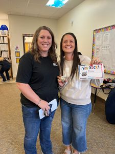 Two women smiling in a classroom, holding a colorful sign and a small gift.