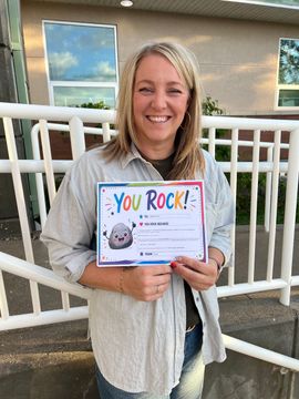 Smiling woman holding a colorful “You Rock!” certificate on a porch with white railing.