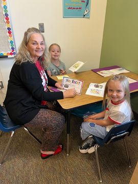 Adult and two children seated at a classroom table, smiling and doing an activity with papers and crayons