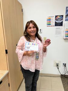 Woman in a pink blouse holding a colorful sign in a classroom or office setting.