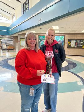 Two people smiling in a school lobby, holding a small sign, with blue-and-white mosaic flooring.