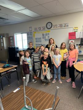 Children posing together in a classroom, with tables, chairs, and a wall clock visible.
