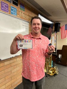 Teacher holding a certificate and trophy in a classroom, smiling beside a whiteboard and school flags.