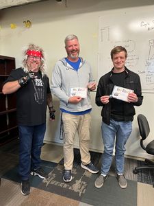 Three people standing indoors, smiling and holding certificates in an office-like room.