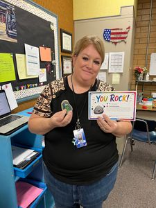 Teacher in classroom holding a “You ROCK!” card and a small award, smiling beside a bulletin board.