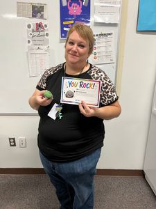 Woman holding a “You Rock!” sign and a small green item, standing in an office or classroom.