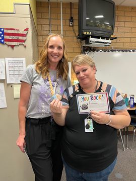 Two women smiling in a classroom, holding a colorful award sign and certificate.
