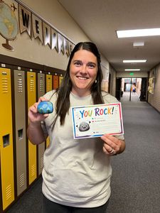 Woman in school hallway holding a colorful ball and a “You rock!” sign, smiling near yellow lockers