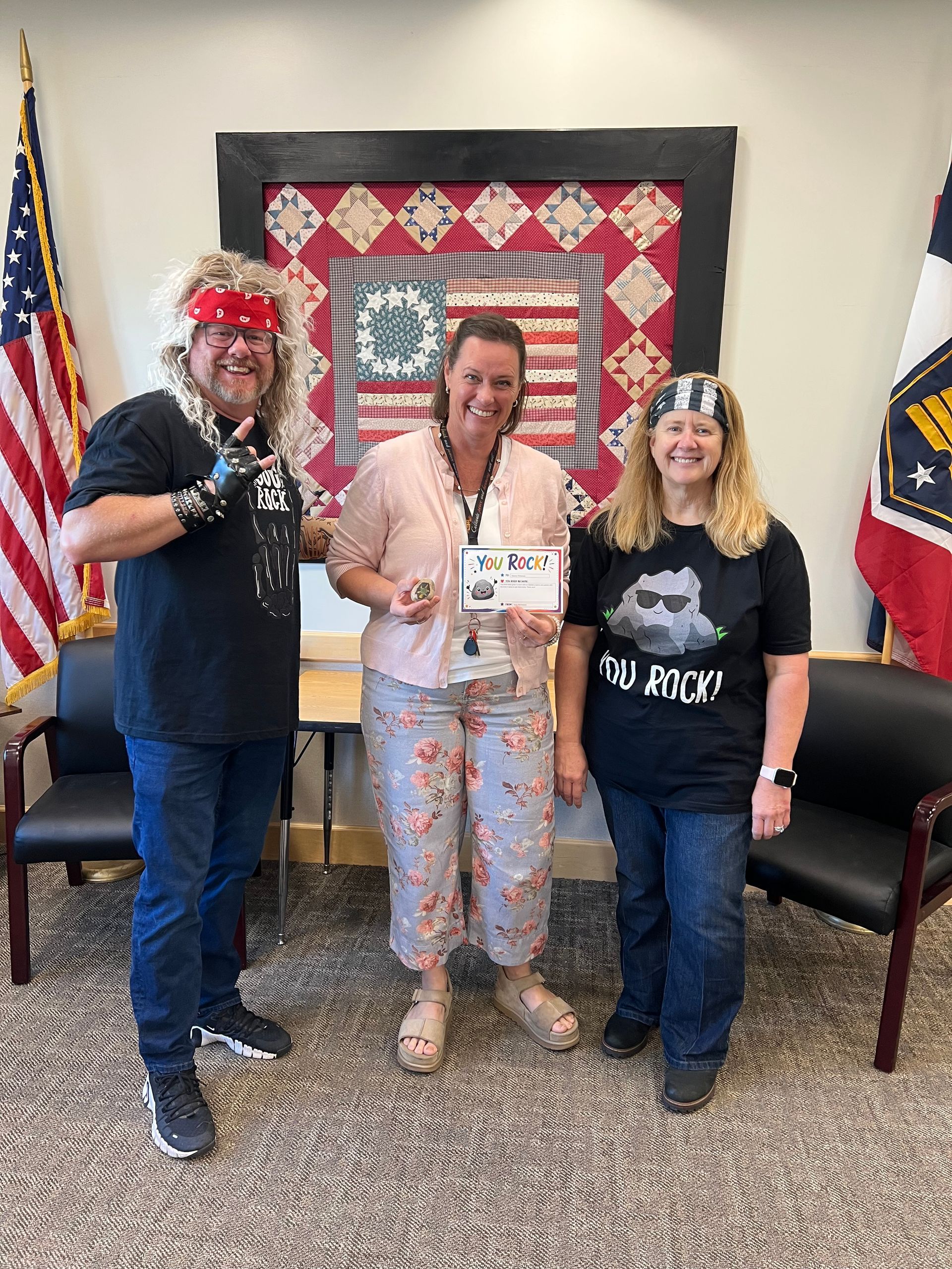 Three people posing indoors, holding a certificate, with U.S. and state flags in the background.