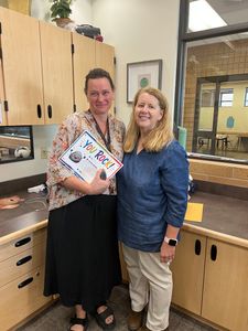 Two women smiling in a classroom, holding a colorful book and standing by wooden cabinets.