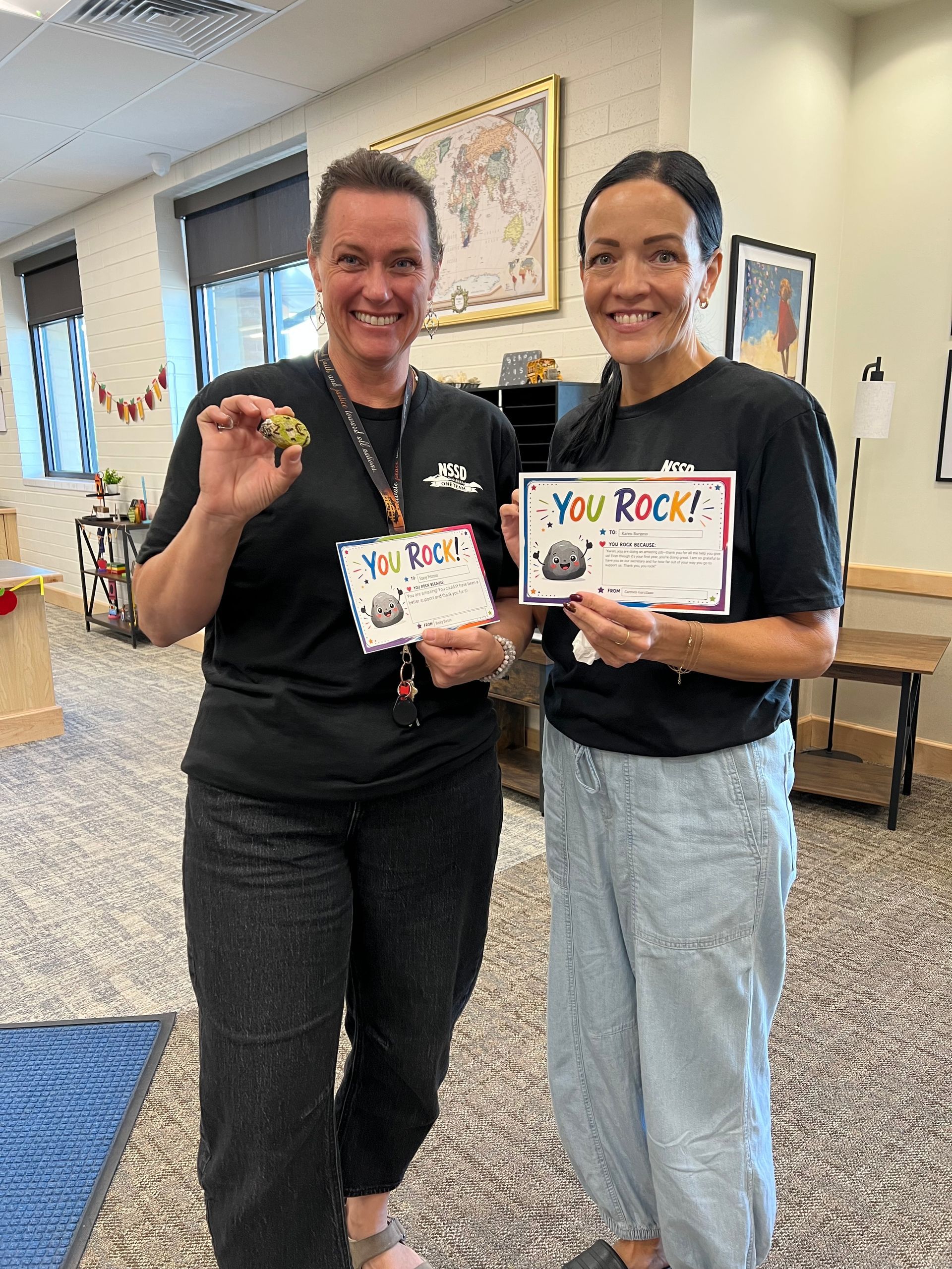 Two people in a room smiling and holding small handmade signs, one reading “You Rock!”