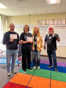 Four people stand on a colorful classroom rug, smiling and holding papers in front of a decorated wall.