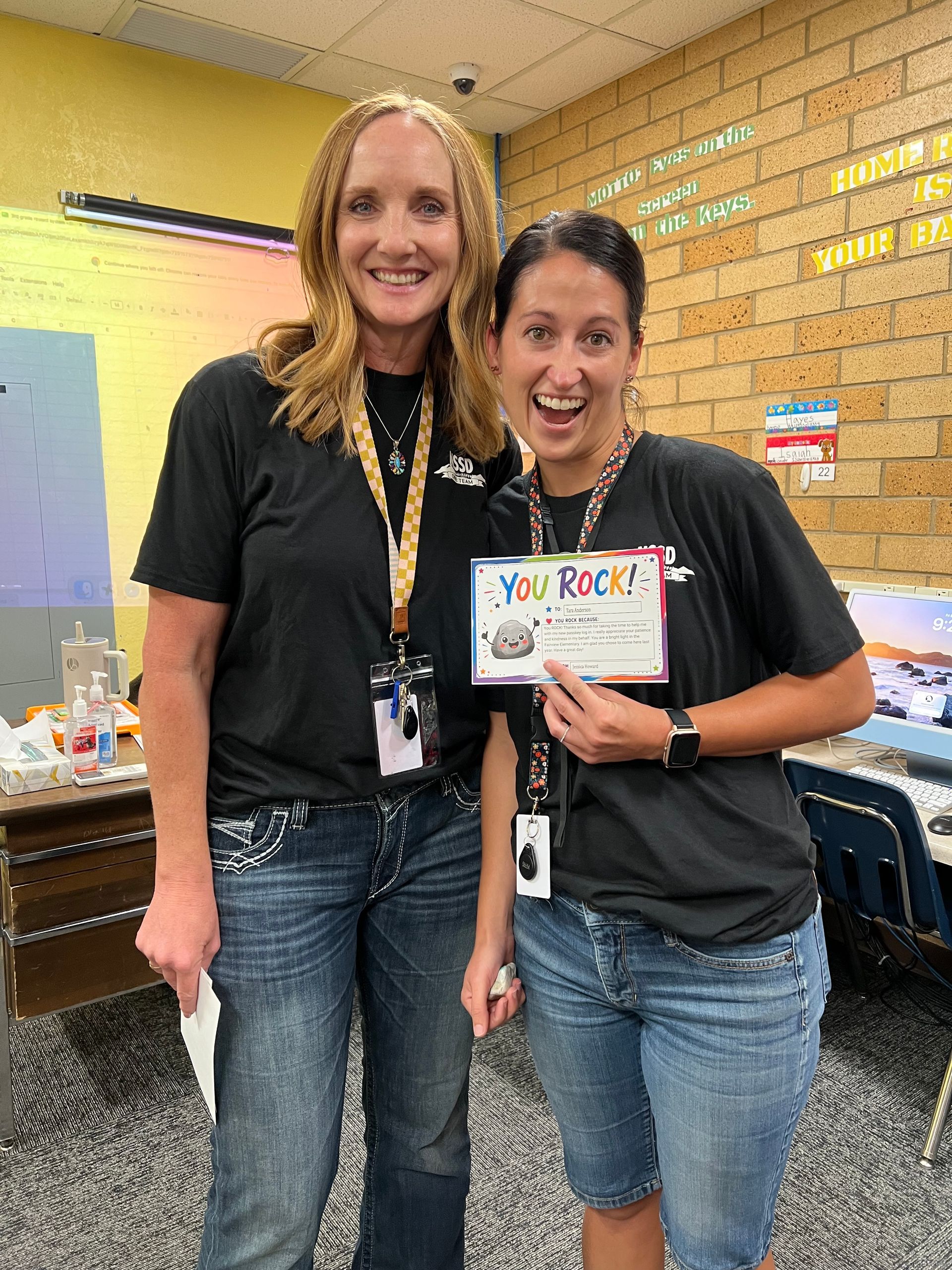 Two women in a school hallway smile while holding a “You ROCK!” sign.