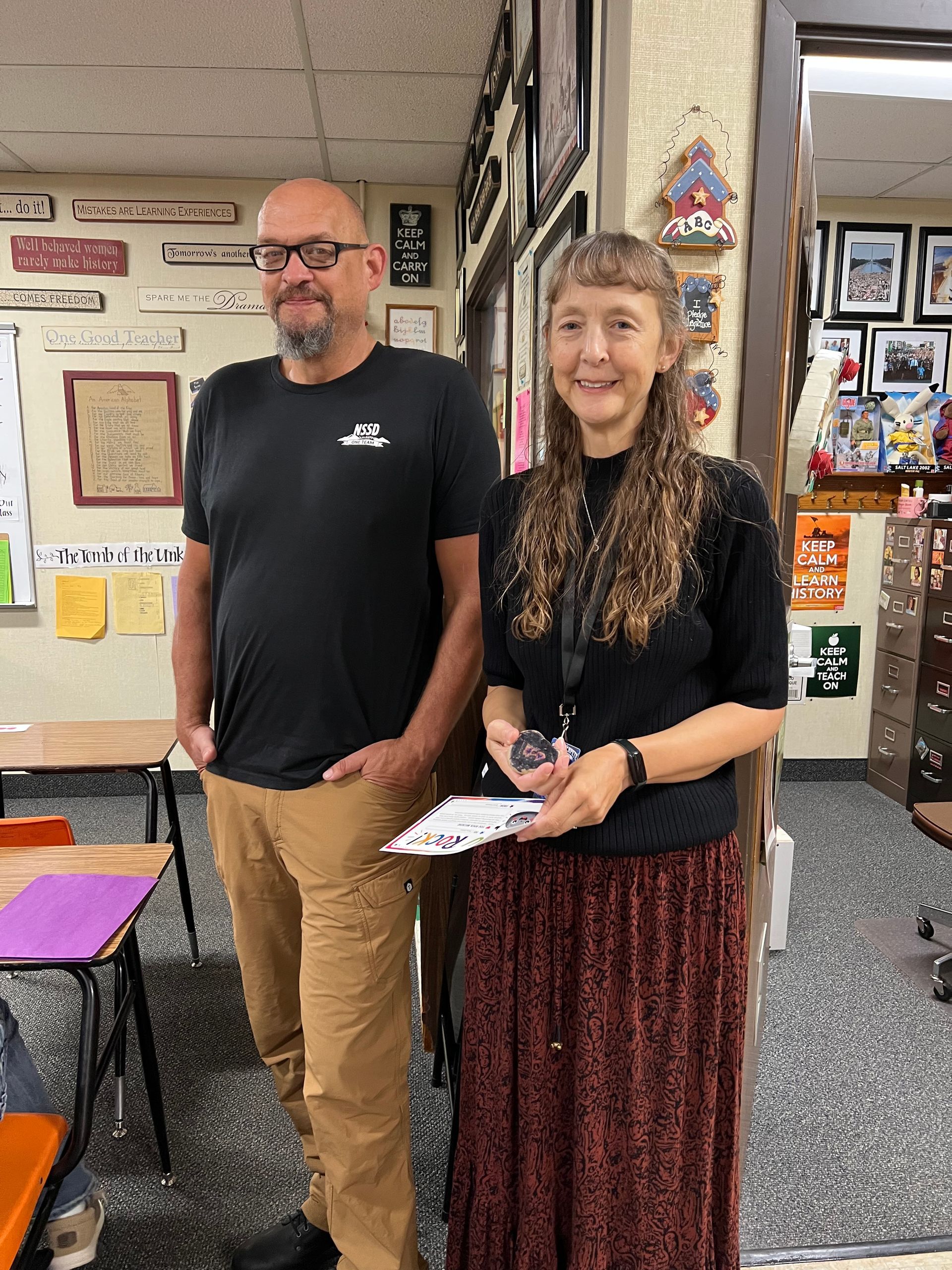 Two people posing indoors, one holding a paper, with colorful wall decorations behind them.
