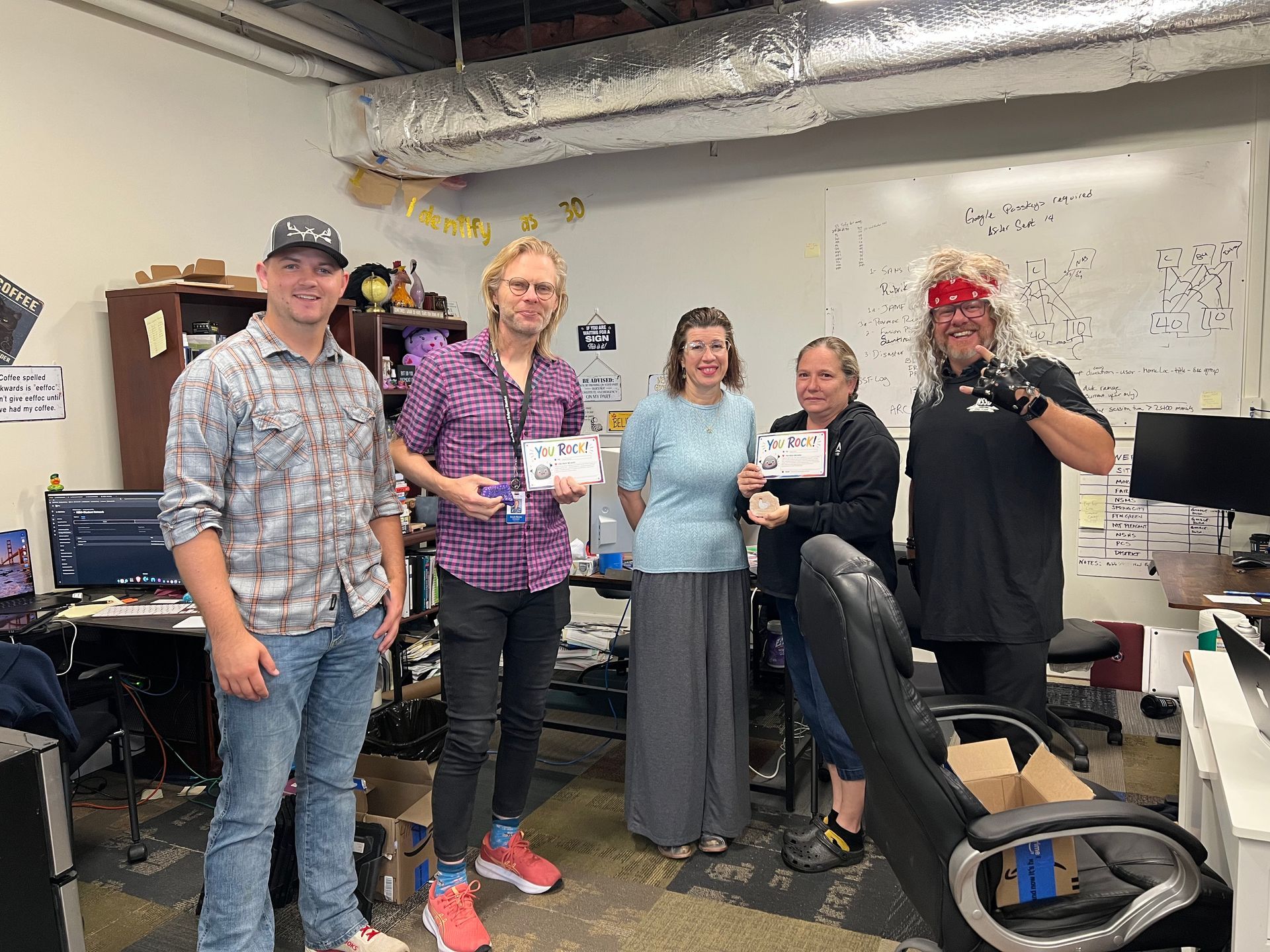 Five people smiling in a workshop, holding coffee mugs near a desk and office equipment