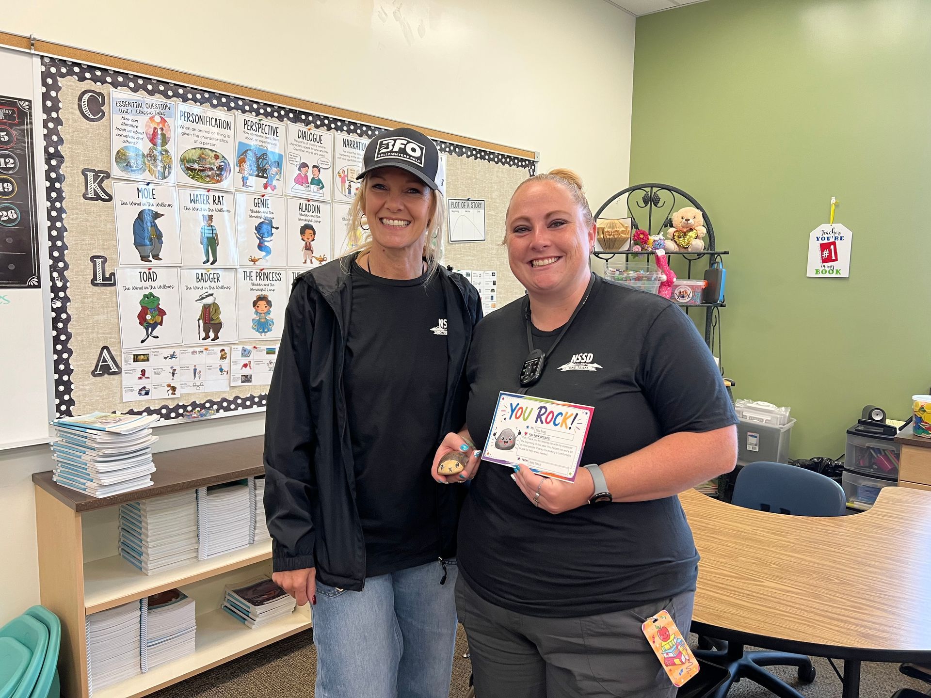 Two smiling people standing in a classroom, one holding a small card in front of a bulletin board.