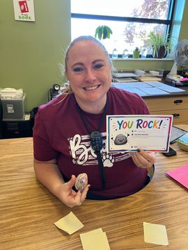 Smiling person holding a “You Rock!” card and treats at a table in an office or classroom