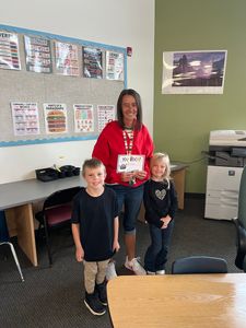 Two children stand with a smiling adult in a classroom beside a desk and bulletin board.