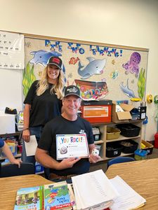 Teacher and student in classroom, smiling beside a colorful wall display and an open book on the desk.