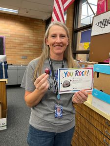 Smiling woman holds a “You ROCK!” sign and a small heart craft in a school hallway