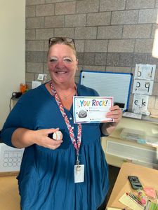 Woman holding a “You Rock!” sign and a small item beside a table of office supplies.