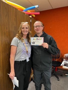 Two smiling people holding a certificate in an office, standing before an orange wall and balloons.