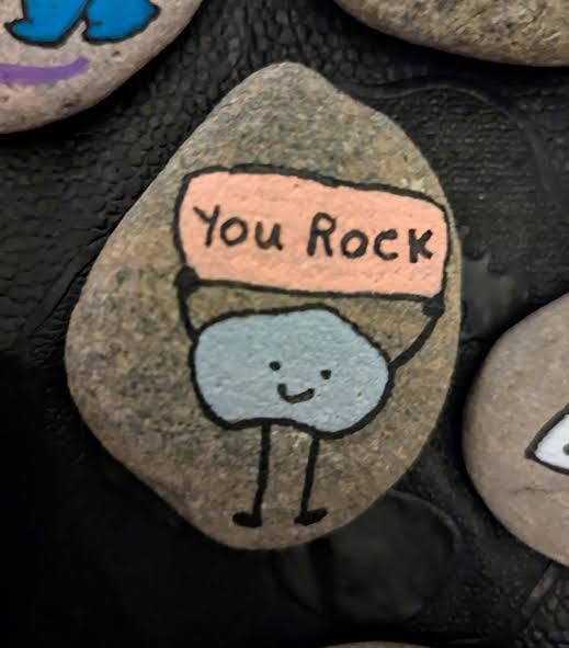 Hand-painted pebble with a smiling figure holding a sign that says “You Rock”