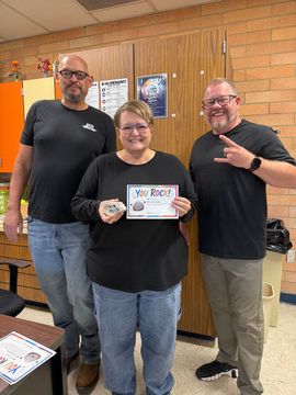 Three people posing indoors, one holding a framed certificate and another making a peace sign.
