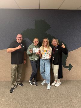 Four people smiling and posing indoors in front of a dark wall mural, making hand gestures