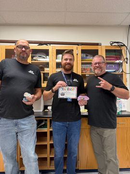 Three men standing in a room, smiling and holding small items in front of wooden cabinets.