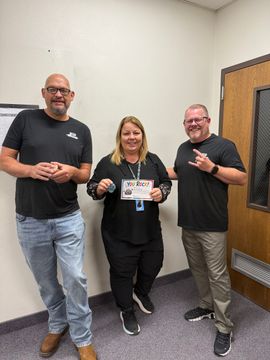 Three people standing in an office, smiling and holding a small sign.