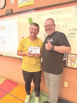 Two people smiling in a classroom, holding a card and giving a thumbs-up in front of a whiteboard.
