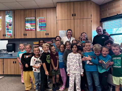 Group of smiling children and two adults posing in a classroom kitchen with cabinets and windows.