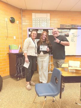 Three people posing in a classroom, smiling and holding an award certificate.