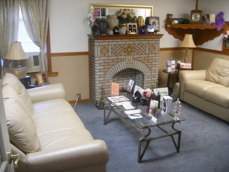 Cozy waiting room with cream-colored couches, brick fireplace, and glass coffee table, possibly in a funeral home.