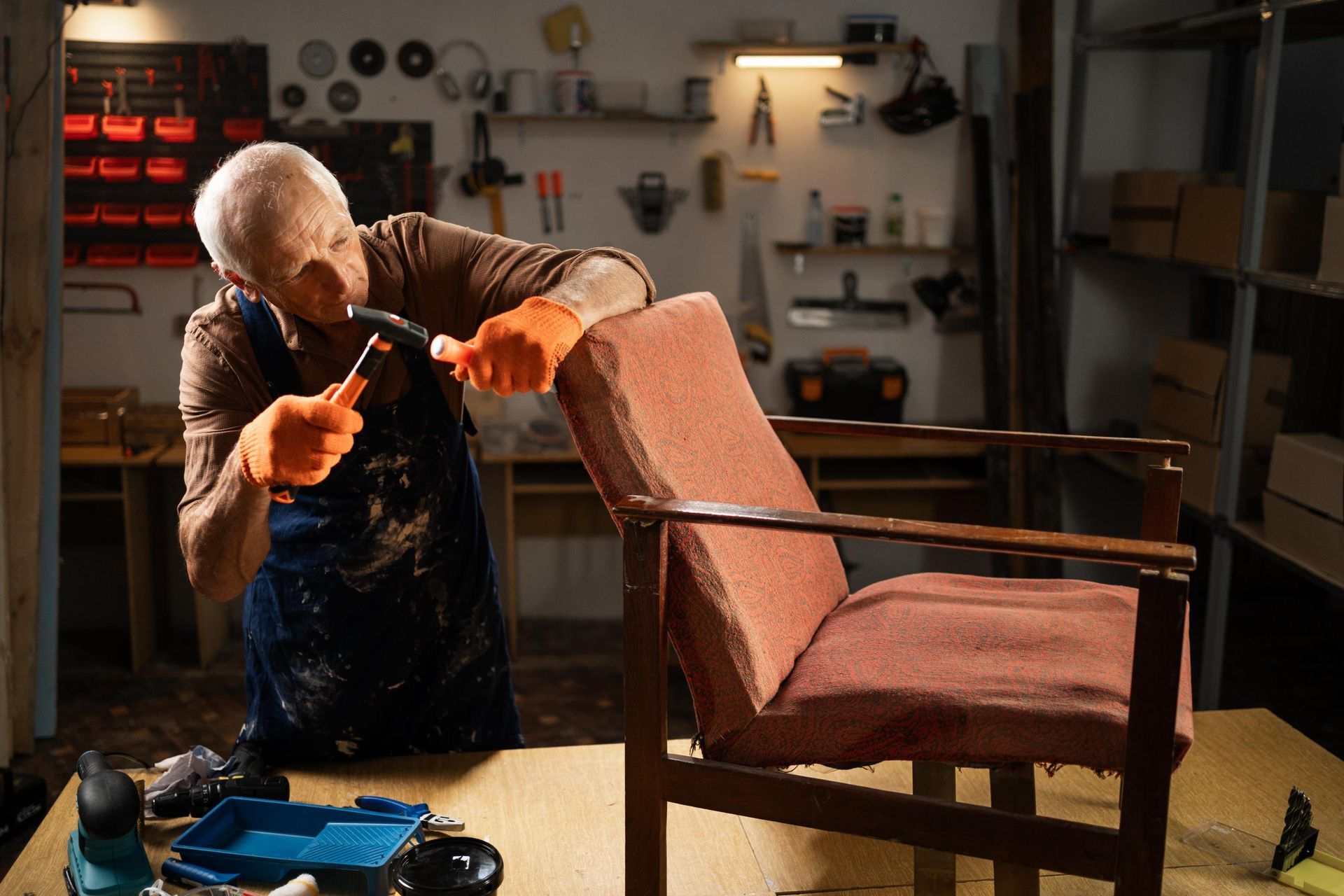 Elderly man in orange gloves repairs a chair in his workshop.
