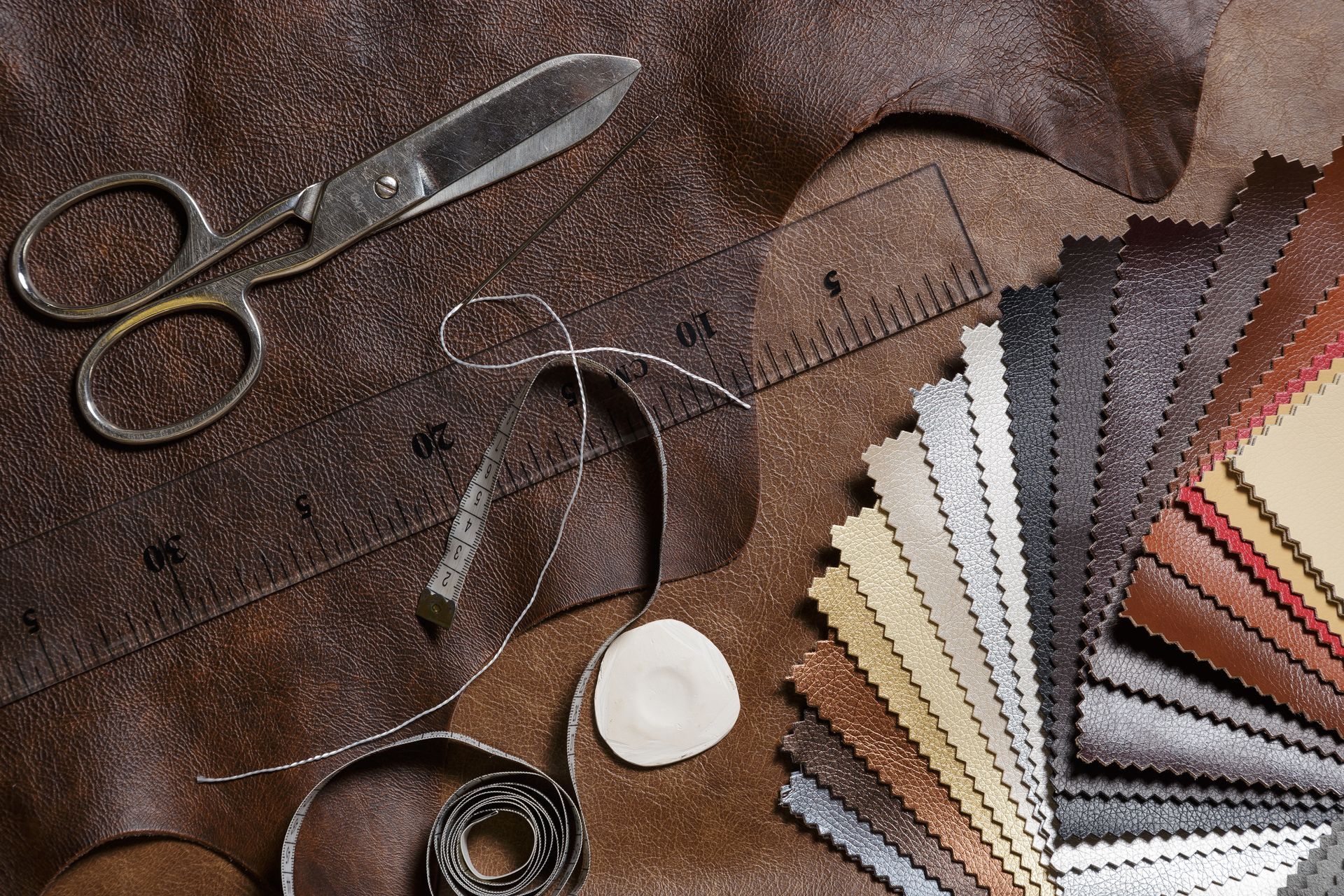 Scissors, a ruler, measuring tape, and fabric swatches in various colors spread across brown textured leather.