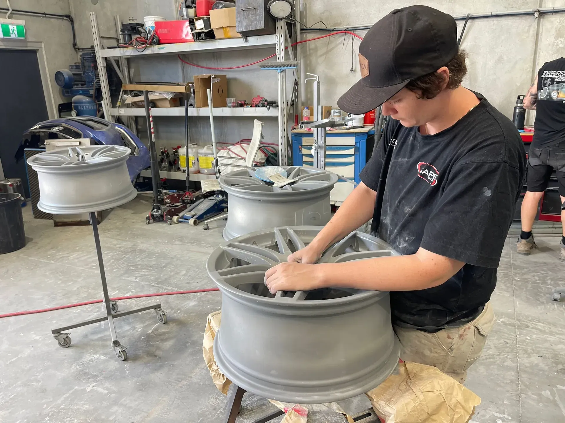 Person working on a car wheel, indoors, workshop. Gray wheels and tools visible. — Coolum Smash Repairs in Coolum Beach, QLD