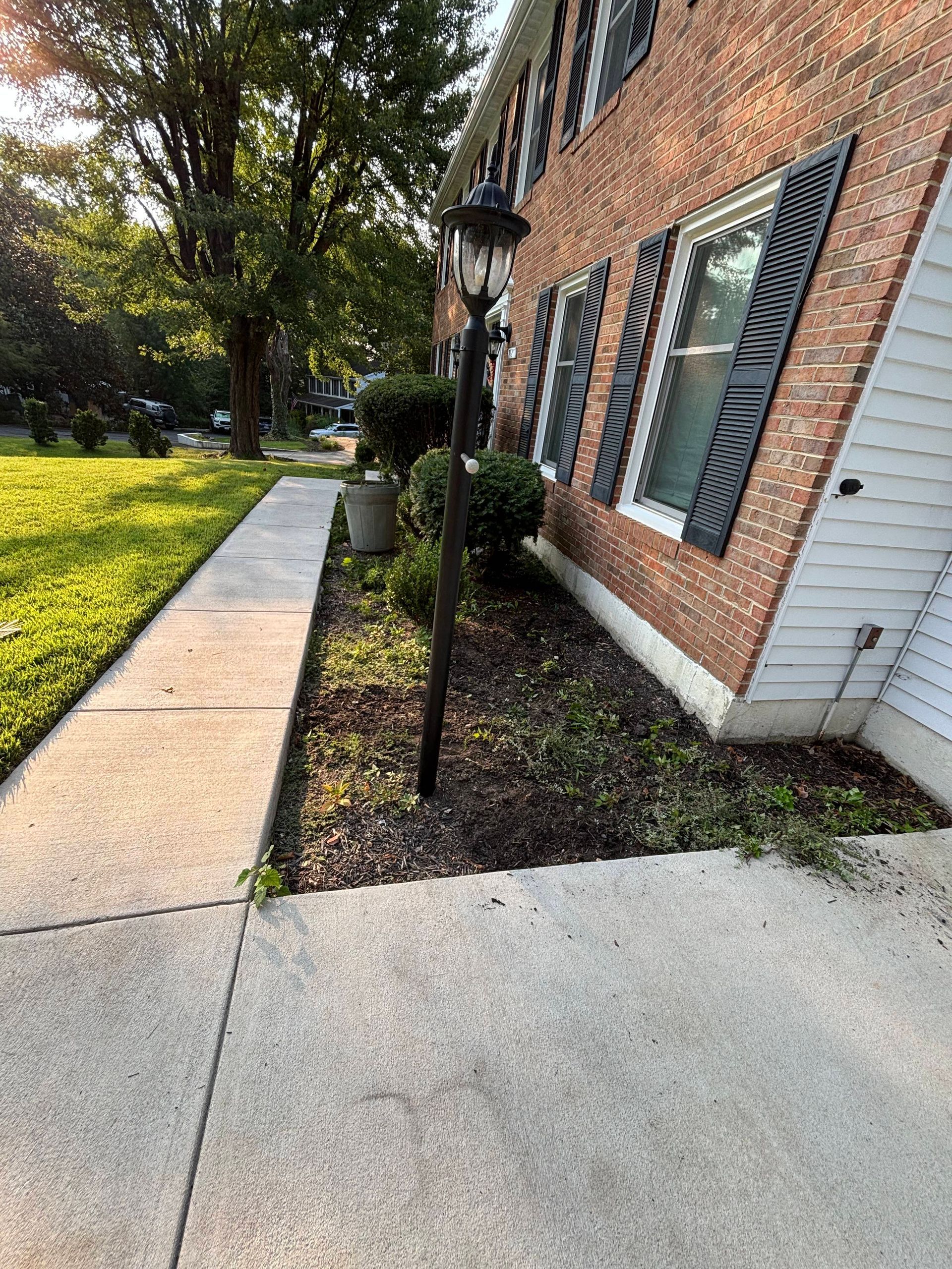 Brick building with green shutters beside a sidewalk, grass, and trees in bright sunlight