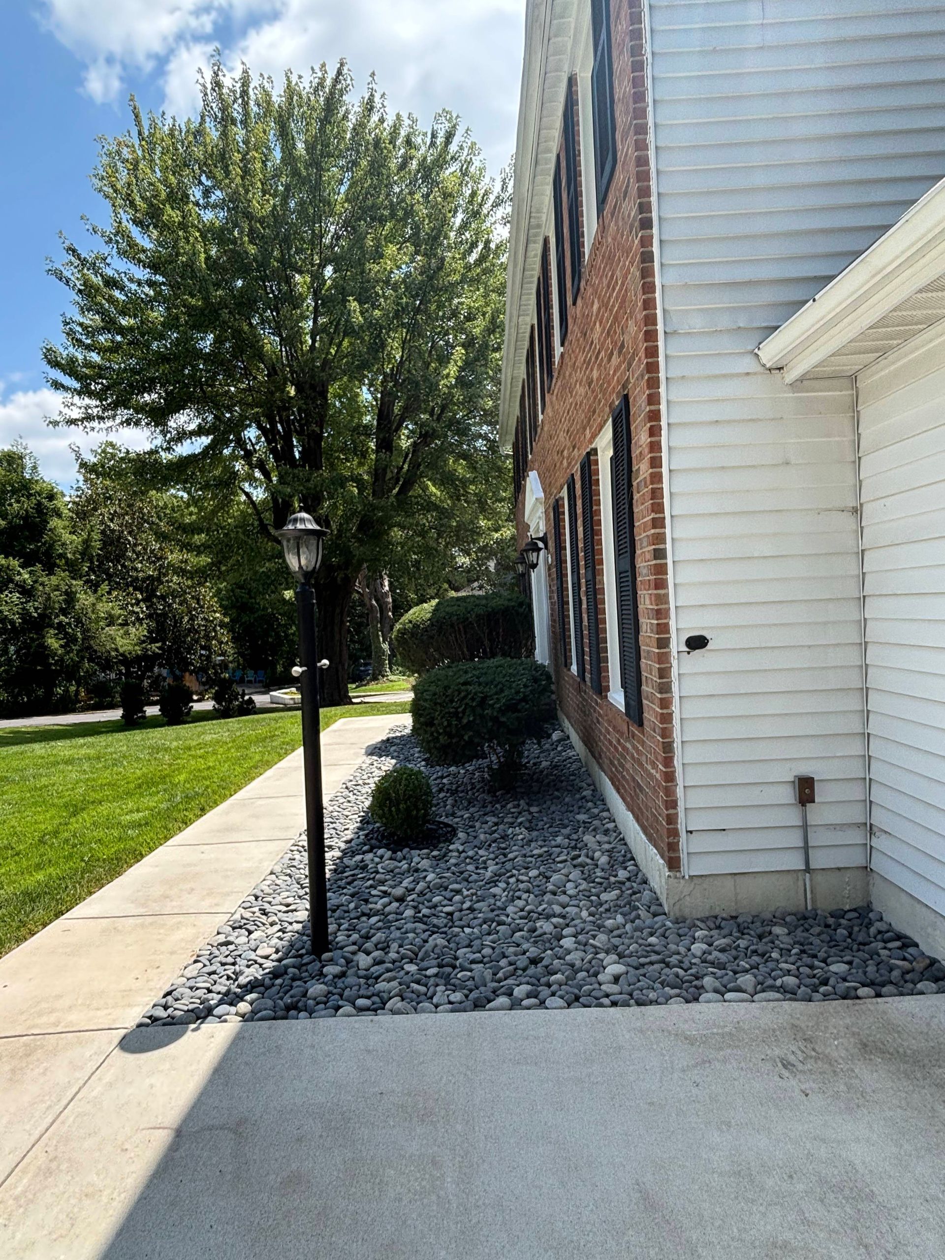 Sidewalk beside a white house with brick siding, gravel landscaping, and a tree along a grassy lawn.