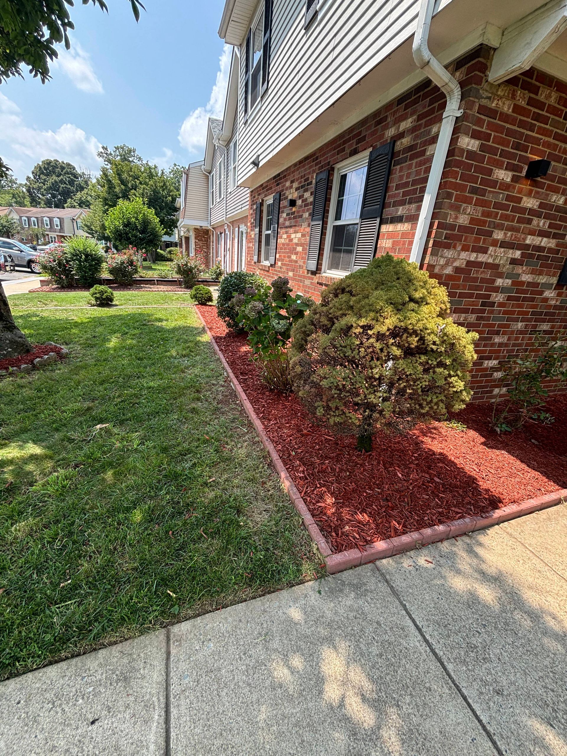 Apartment exterior with brick siding, green lawn, red mulch beds, and concrete walkway under a bright sky