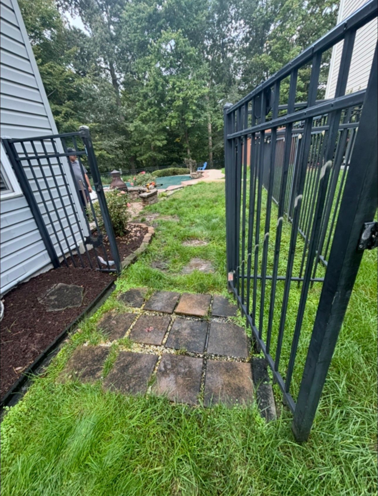 Open black metal gate beside a house, leading to a grassy yard with stone pavers and trees beyond.
