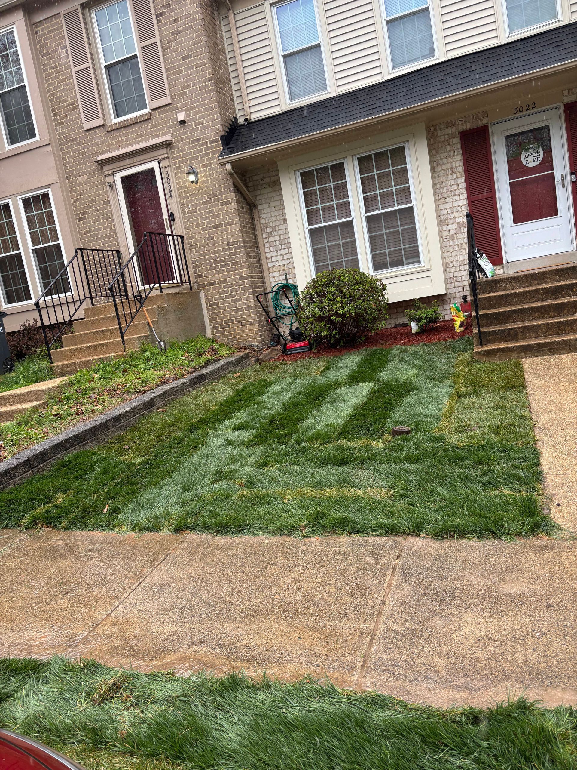 Suburban townhouse row with fresh green lawn, brick facades, red doors, and a wet sidewalk in front.