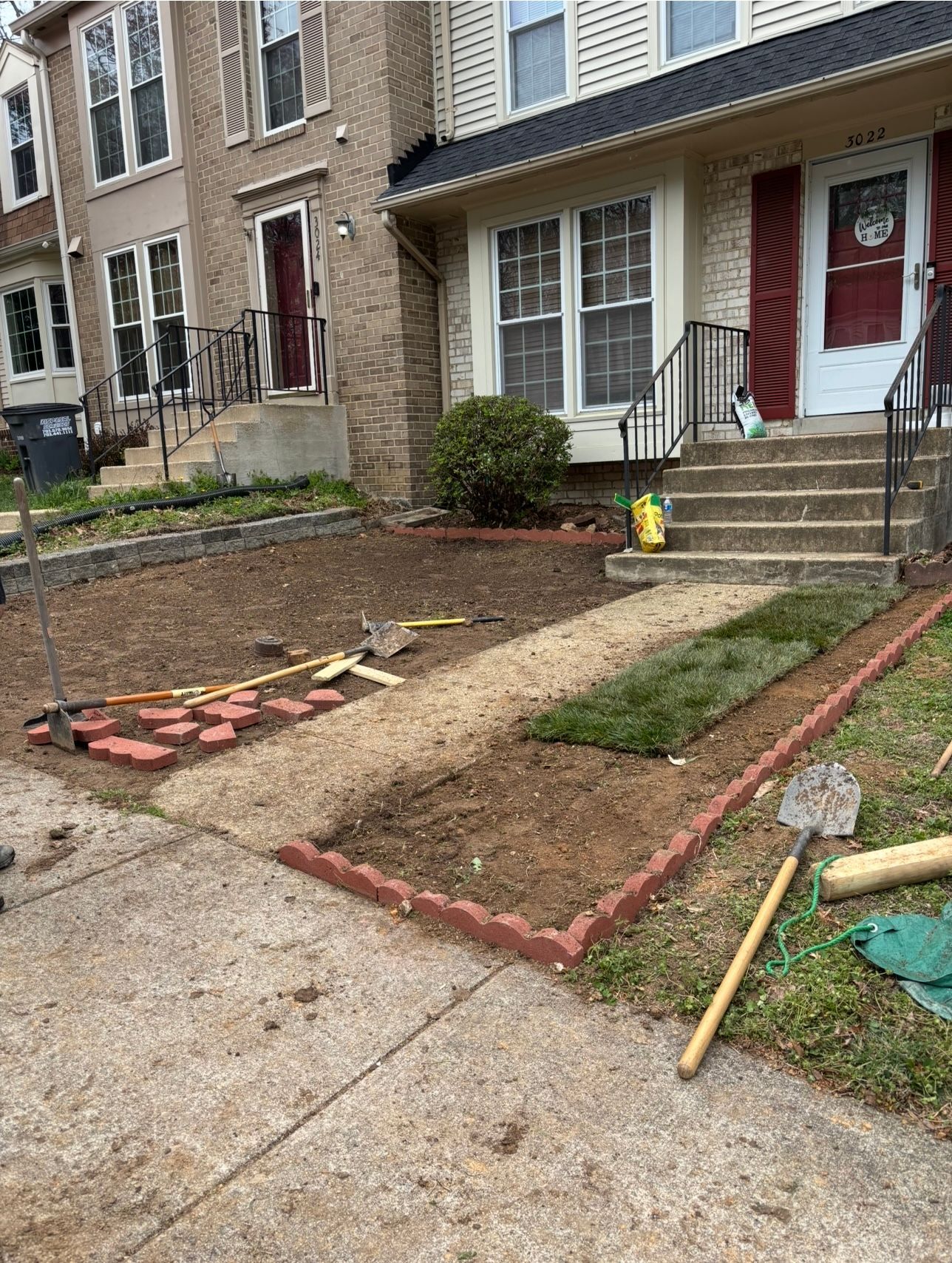 Front yard under renovation with bare soil, red edging, and tools beside townhouse steps