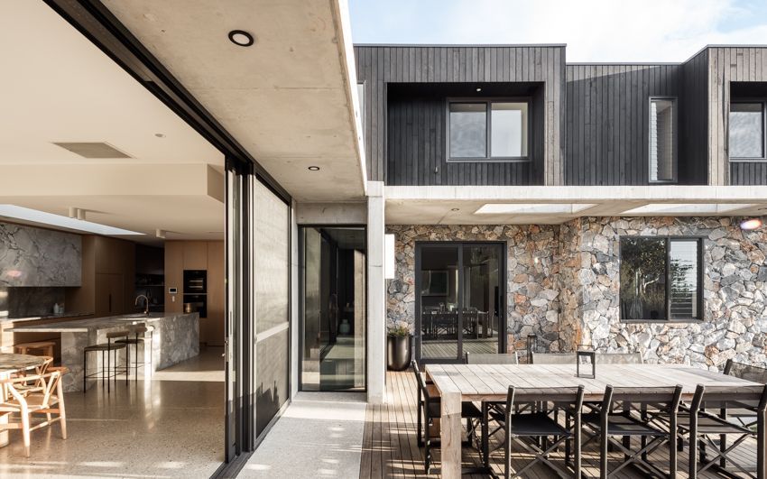 Outdoor patio with dining table, adjacent to a kitchen with open sliding doors. Modern house with stone and dark wood siding — Make The Mark Building Services in Illawarra, NSW