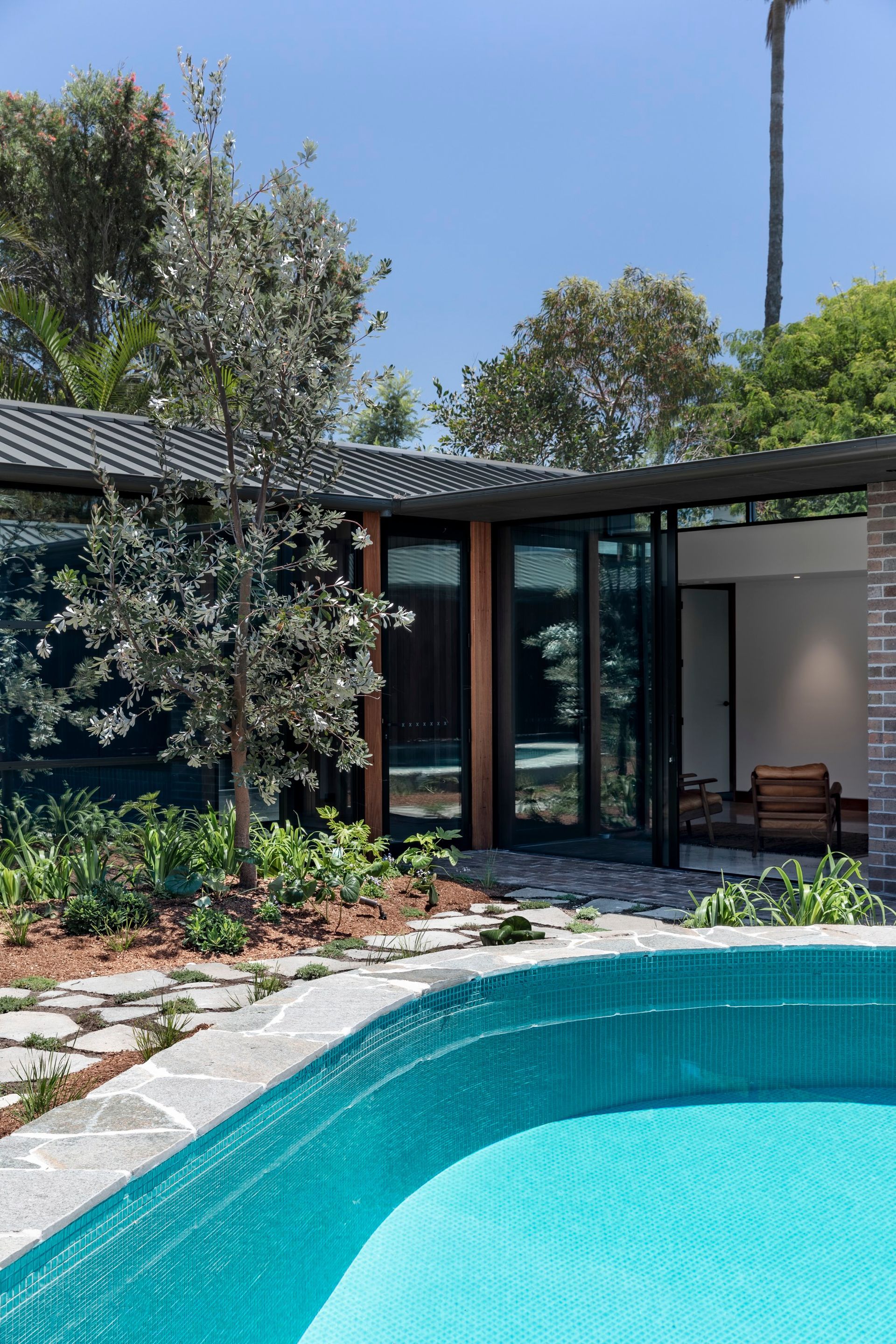 Poolside view with turquoise water, a house with open doors, and landscaping. Blue sky — Make The Mark Building Services in Illawarra, NSW