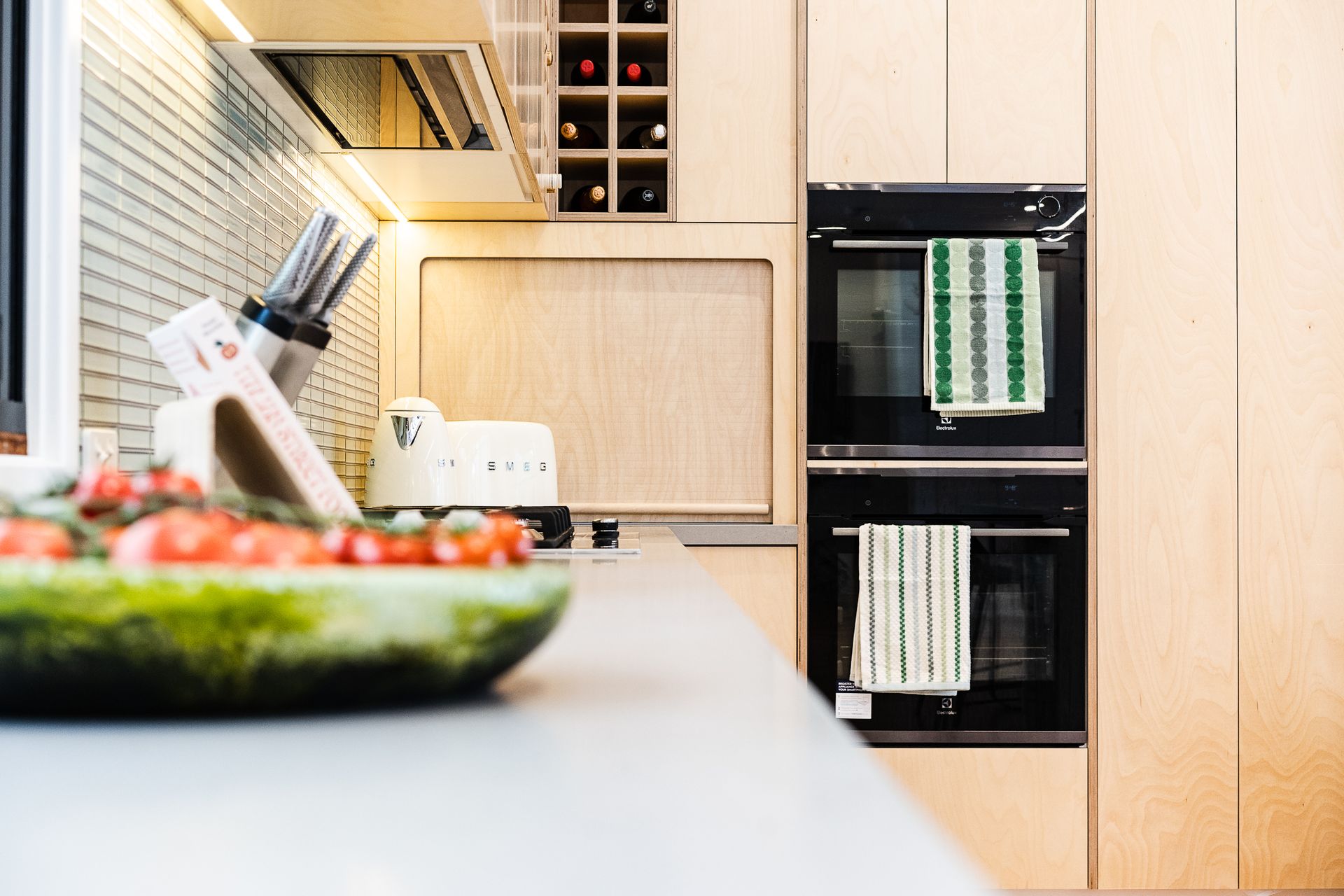 Kitchen with light-colored cabinets, double ovens with towels, a counter with a bowl of food, and a wine rack - Make The Mark Building Services in Mangerton, NSW