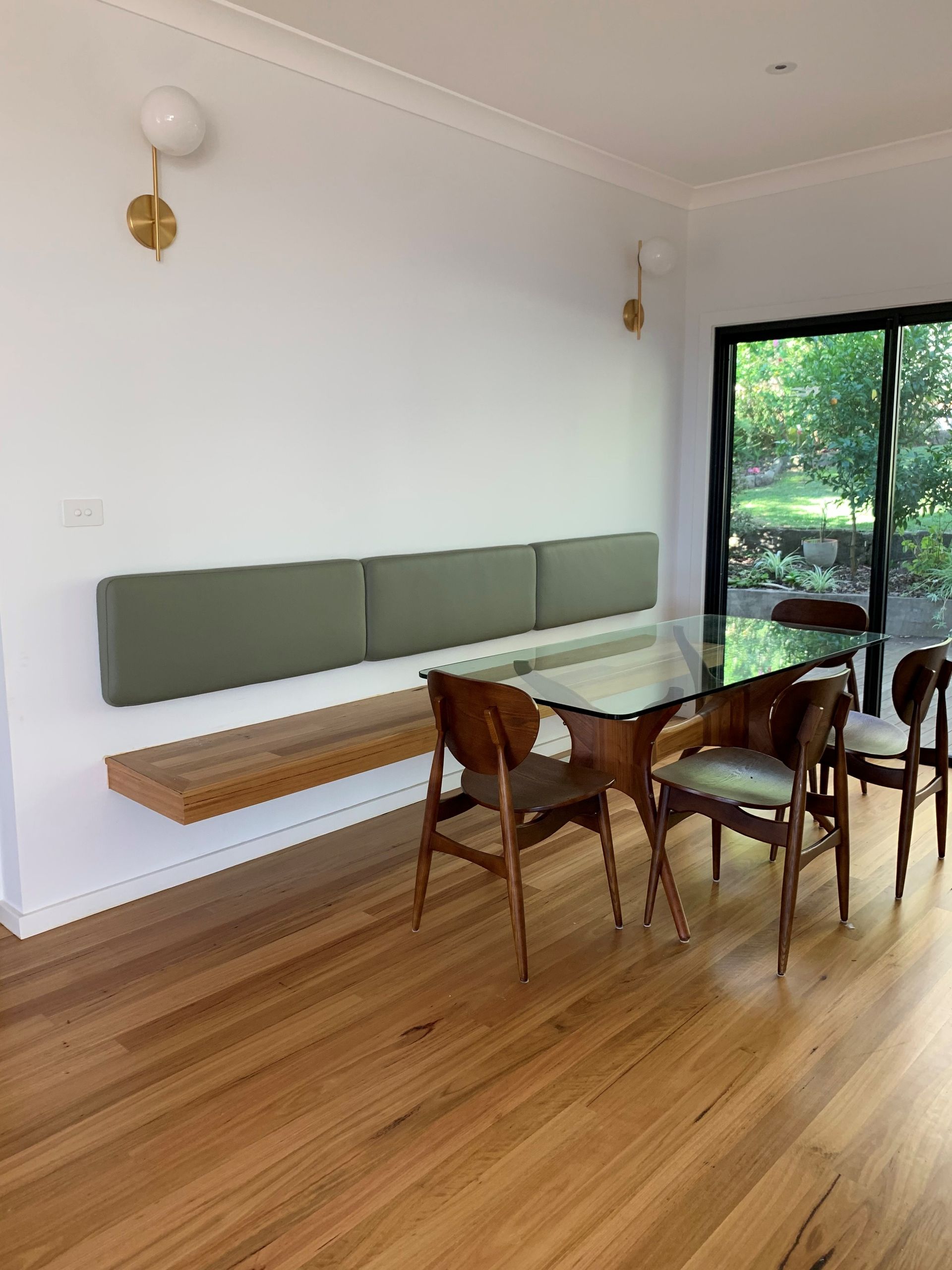 Dining area: floating wood bench, green padded backrest, glass table, wood chairs, brass sconces, hardwood floor - Make The Mark Building Services in Mangerton, NSW