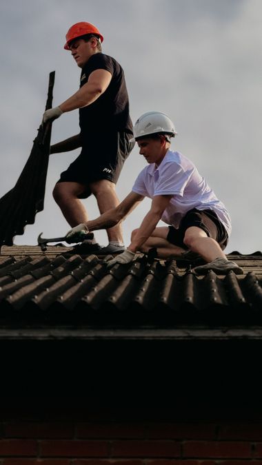 Two workers in hard hats roofing a house, one standing with a tool and one crouching on the shingles.