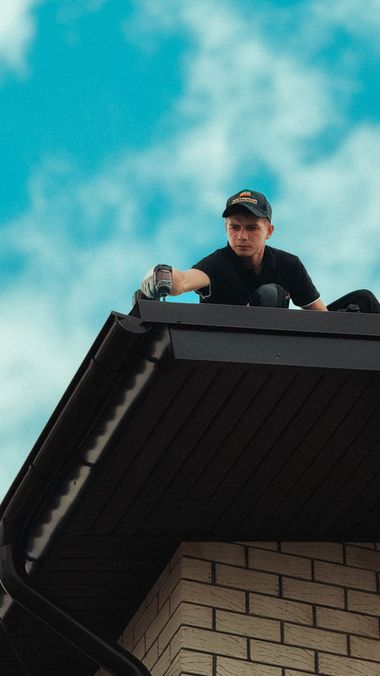Person on a roof, wearing a cap, painting or repairing the eaves against a blue sky.